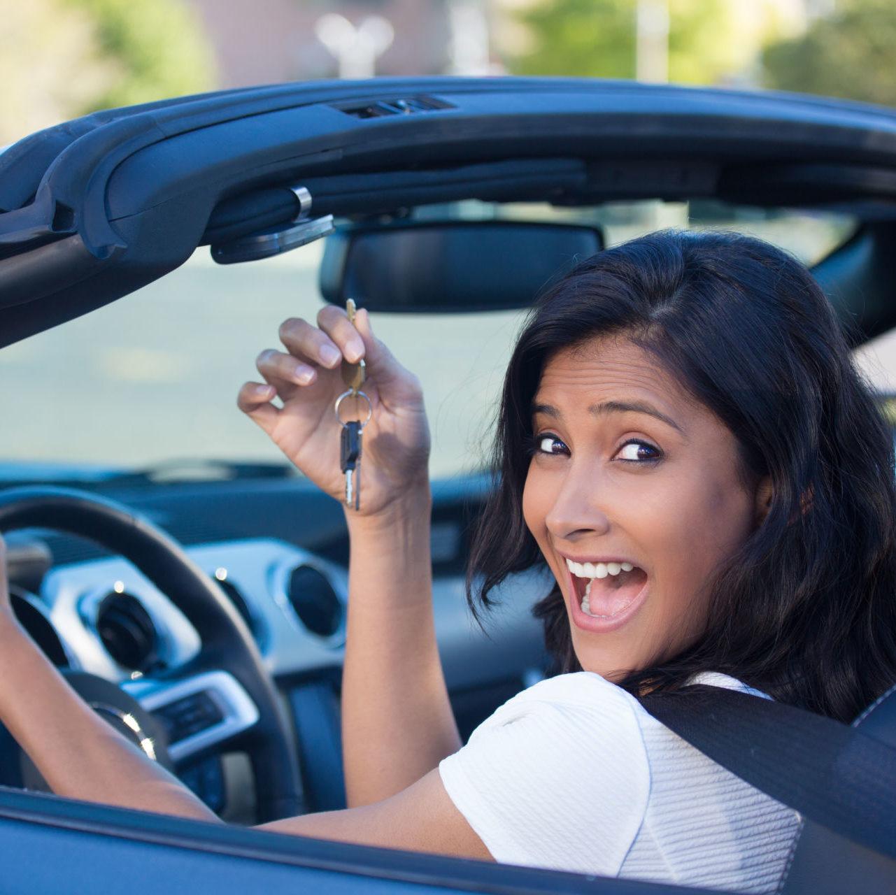 A young, cheerful woman holding up keys to her first new sports car