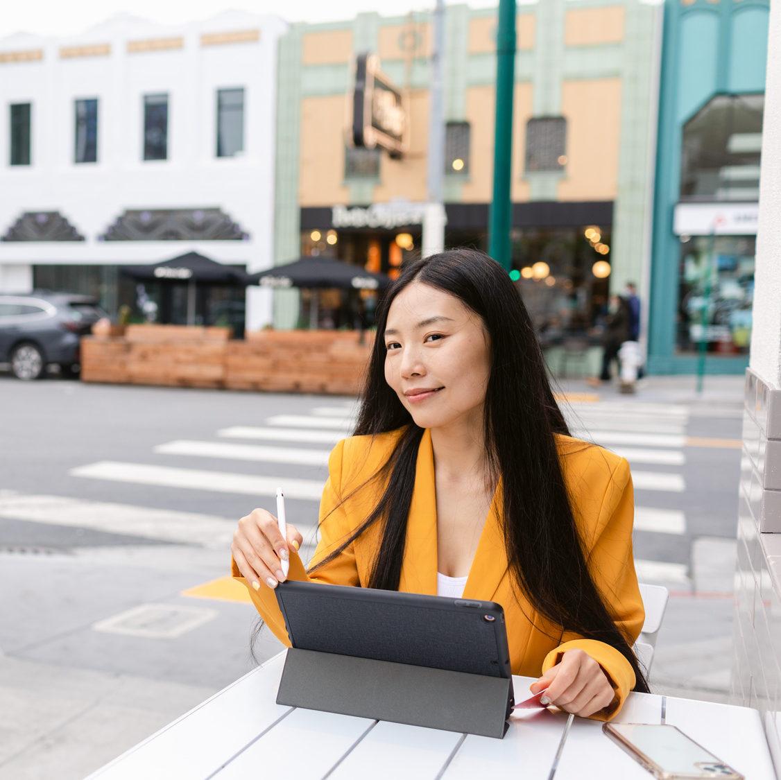 Young Chinese female with credit card and tablet looking away and thinking about online purchases while sitting at table in street cafeteria in Bay Area