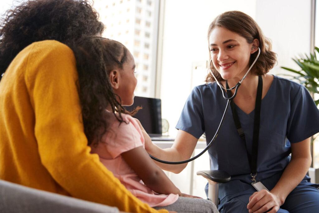 A mom with her daughter sitting in her lap and a doctor using a stethoscope to check the daughter's heartbeat