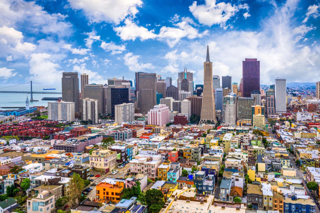 A panoramic view of the San Francisco skyline shows downtown skyscrapers, including the Transamerica Pyramid, with the Bay Bridge and waterfront visible in the background.