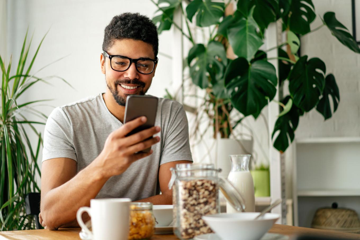 A man wearing glasses smiles while looking at his smartphone at a breakfast table with coffee, cereal, and milk in a bright kitchen with houseplants.