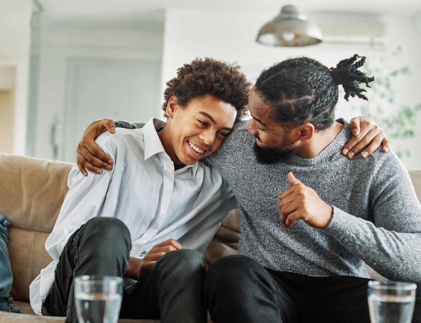 A father and teenage son sit close together on a couch, smiling and sharing a warm, affectionate moment in a cozy living room.