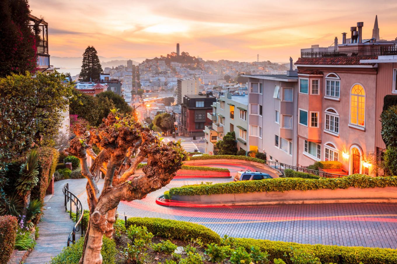 A scenic view of Lombard Street’s winding curves in San Francisco at sunset, with city buildings and Coit Tower visible in the background.